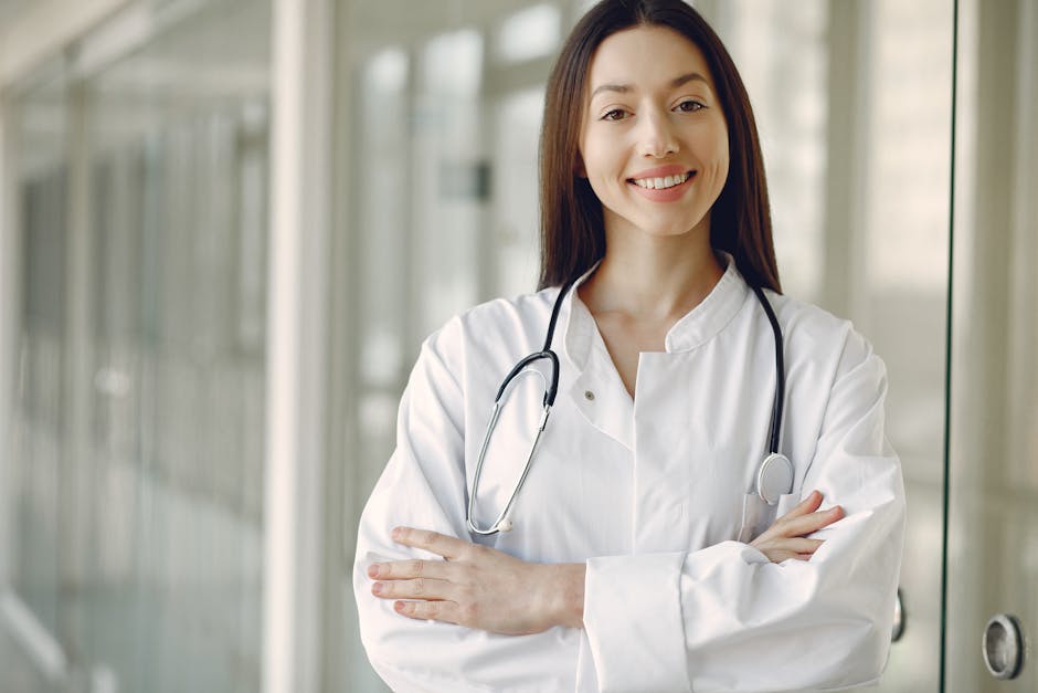 Crop cheerful ethnic female doctor in medical uniform and stethoscope holding hands crossed standing in modern clinic corridor